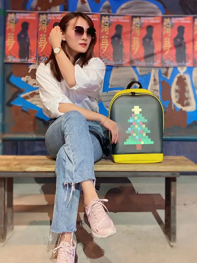 Stylish individual wearing sunglasses, sitting with Gelrova T2 LED Backpack displaying a pixelated Christmas tree design.