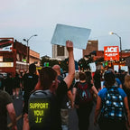 Supportive crowd at a protest, with a person holding a sign and wearing a shirt that reads 'SUPPORT YOU! J.r'.